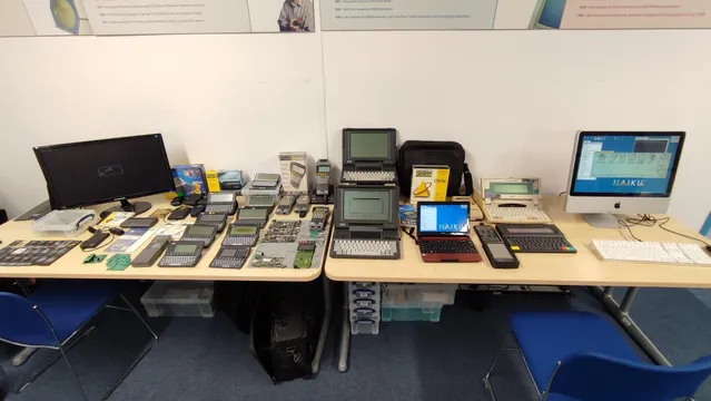 A photo of my stand at the Festival of Portable Computing (early 2024) at the Centre for Computing History, Cambridge, UK. On the table is a selection of Psion devices, including various SIBO/EPOC16 devices (Series 3/3a/3c/3mx, Workabout, Siena, MC400), a few EPOC32 devices (Series 5/5mx), a few PsiDrive boards, many SIBO SSDs, some boxed software. There is also an Amstrad NC100, a Microwriter MW4, an Acer netbook and a 2009 iMac. The Acer and iMac are running Haiku. The iMac is running the MAME-based Psion SIBO emulator.