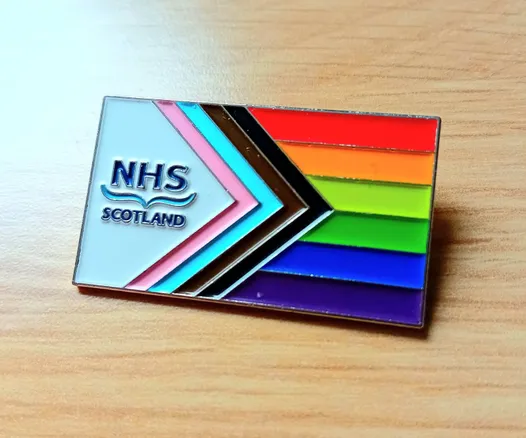 Metal pin badge sitting on a wooden desk, featuring the Progress Pride flag: the white space to the left is extended and is occupied by the NHS Scotland logo.