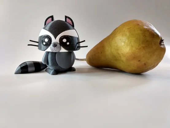 A multi-colored 3D-printed cartoon raccoon standing to the left of a pear lying on its side on a white background.