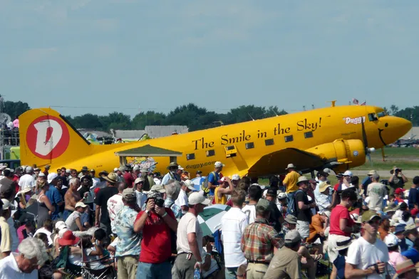 "A large crowd is gathered at an airshow, focusing on a bright yellow airplane with 'The Smile in the Sky!' and 'Duggy' written on its side. The airplane has a smiley face painted on its nose and a large red 'D' on its tail. The website 'duggy.com' is also visible on the side of the plane. Many people are taking photos and observing the aircraft. The airplane is a vintage model, adding to the event's interest and relevance."