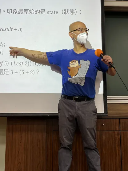 I'm a bald Asian man, standing in front of a computer projector screen, wearing gray slacks and a blue T-shirt that pictures a boot and an otter. I'm holding a microphone with my left hand and pointing at text on the screen with my right hand. The text is a mix of math, English, and Chinese. I'm masked, wearing glasses, and looking to the side.