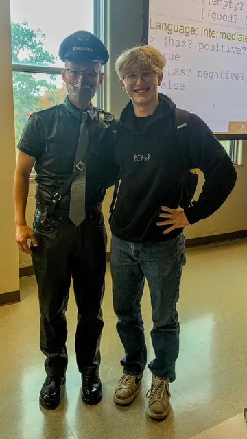 I'm an Asian man wearing a brimmed uniform hat that says "conductor", leather shirt, leather tie, Sam Browne belt, leather pants, and Corcoran boots with a shiny toe. I'm standing next to a smiling young white man in hoodie, jeans, and sneakers. We're standing in a classroom in front of a bright window and projected computer programming screen.