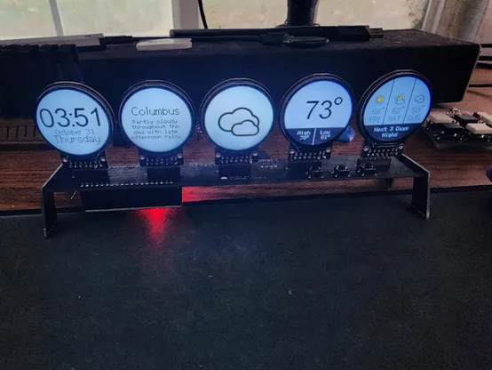 Photo of a set of 5 circular displays connected to a pcb with an esp32 at the bottom. The displays show a clock the weather described in text, an image of the weather (in this case clouds), the current high and low temps, and the next 3 day forecast