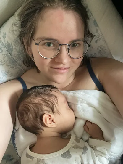 A nonbinary person with light brown hair and glasses slightly smiles up at the camera. They have a sleeping dark-haired baby on their chest.