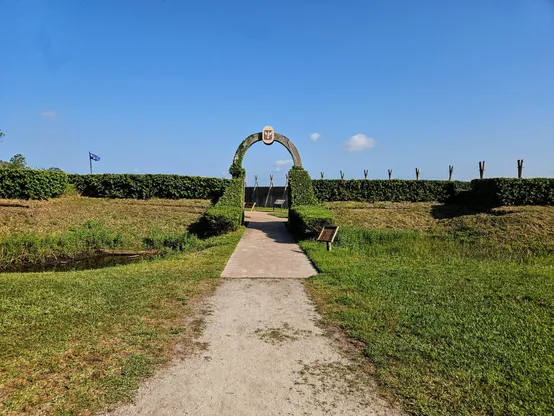 Approaching the exhibit for Fort Caroline's Fort, at the end of a long well worn pathway, a large area is surrounded by thick green hedges, with an arched entrance.