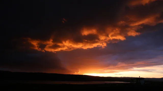 A dramatic sunset above a river. Leading south-east, light shafts break into dark looming clouds, while to the north, blue clouds with tan and peach highlights give way to a a pale yellow sky. A bank of nearby orange clouds frame the scene from above