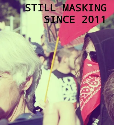 Person at Occupy Wall Street with a black hoodie with a red bandana over their face holding a red flag with the text "Still masking / since 2011"