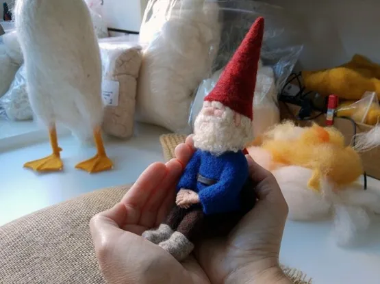 A small needle felted utterly relaxed gnome sitting in two cupped hands, with some needlefelting supplies in the background