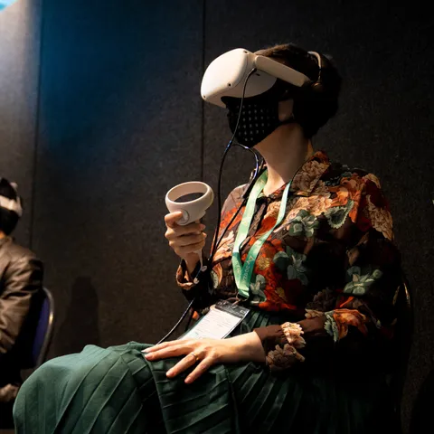 A photograph of a woman with short dark hair, sitting and using a white Virtual Reality headset and controller. She is wearing a checkered face mask, a loud floral shirt and a long teal skirt, as well as a convention lanyard around her neck. 