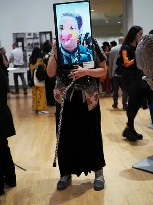 A person faces the camera standing in a crowded art exhibition. They are wearing a screen that displays their face layered with a digital mask. 