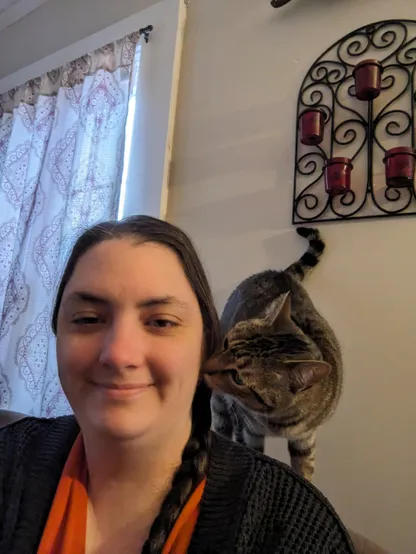 Picture of a white woman smiling at the camera while a cat perches on her shoulder sniffing her face