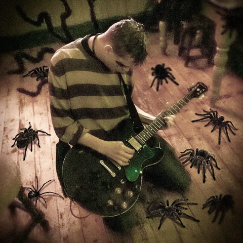 Man kneels on a wooden floor leaning slightly backward with a guitar on his lap. He looks down at his hands playing it. He wears a brown striped top and has black around his eyes. There are giant spiders crawling around him.