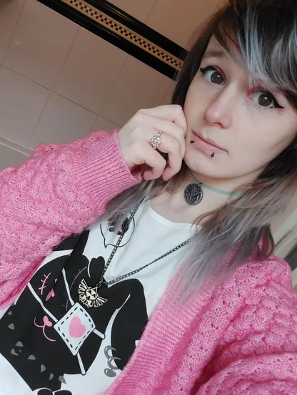 A nonbinary person. They have shoulder length hair which is different colours throughout.
A pink cardigan over a white top with a punk looking rabbit on.