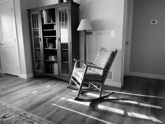 An antique rocking chair sits in the foreground. Behind is a bookcase full of books (some of them banned) and a floor-standing reading lamp. The photo is in black and white.  The chair has been in my wife's family for close to eighty years and was traded to my wife and I by her grandfather for a bottle of his favorite scotch. I think we got the better of the deal. 