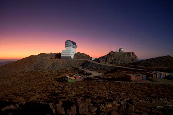 A white observatory building sits atop a brown rocky summit with a small silver dome on a nearby hill. The sky is glowing with beautiful yellow, pink, and purple twilight hues.