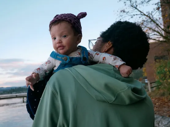 A Black man has his back to the camera, and holds an infant wearing overalls and a hat with ears. The baby is facing to the forward-left and has their arms off to each side.