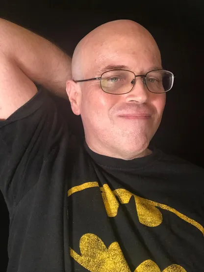 Photo of a white man with shaved head wearing glasses and a slight smile. Has a t-shirt with a Batman symbol on it. 