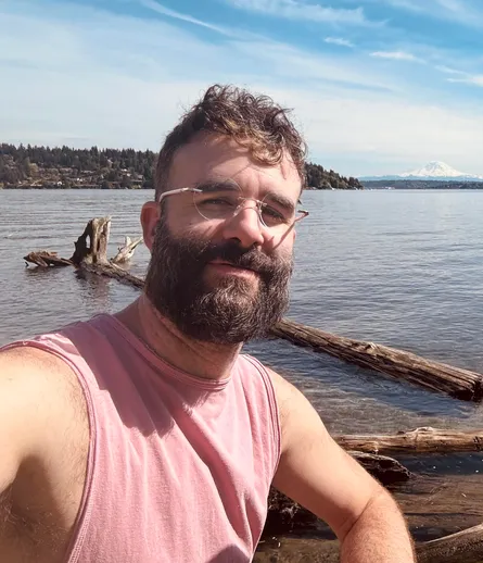 Bearded person wearing a pink sleeveless shirt and circular rimless glasses on the shore of x̌ačuʔ (Lake Washington) with ɬagʷic̓atəb (Mercer Island) and təq̓ʷubəʔ (Mt Rainier) visible in the background.