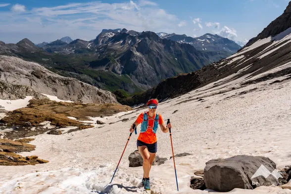 Trail running woman who's wearing an orange t-shirt, shorts and a read hairband is crossing a snow field in the middle of summer in the Swiss Alps.