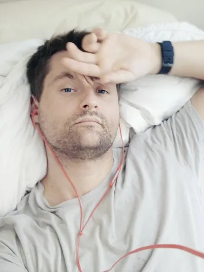 A man taking a selfie of himself with his left hand on his forehead in a lazy afteroon, lying on bed, listening music with hearbuds