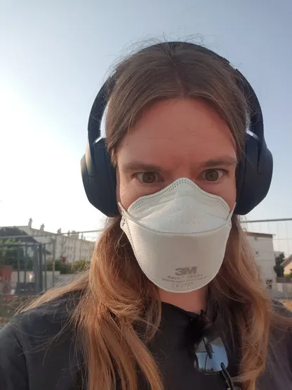 Selfie masqué. Je suis un homme blanc, maigre, aux longs cheveux blonds sous les omoplates. Je regarde l'objectif d'un air insistant caricatural. Mon visage est en partie masqué par un FFP2 Aura, et je porte un casque à réduction de bruit/