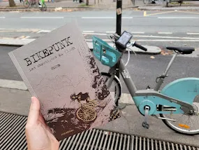 Une main présente la couverture du livre Bikepunk de Ploum devant un vélib électrique bleu accroché à un poteau.