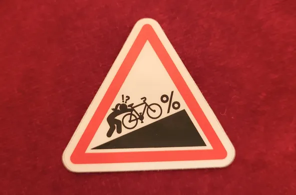 Sticker en forme de panneau de signalisation. Une silhouette de femme poussant un vélo dans une montée à x %