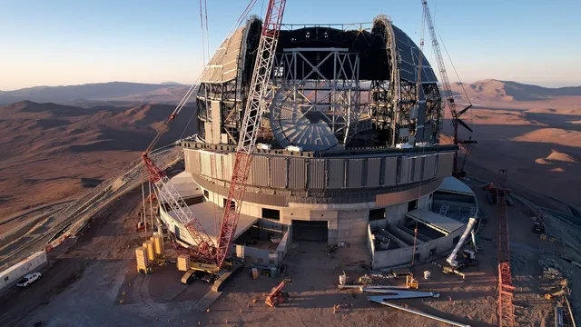 The giant metal frame of a dome takes up most of the picture frame, contrasting with the brown hills and a thin horizontal line of blue sky in the background. The dome's frame is not yet fully covered in the middle, so the dark brown landscape behind it shows. The big opening in the dome's roof shows the interior, still a construction site with a structure of white beams. In front of the big building is one red crane, taller than the dome.