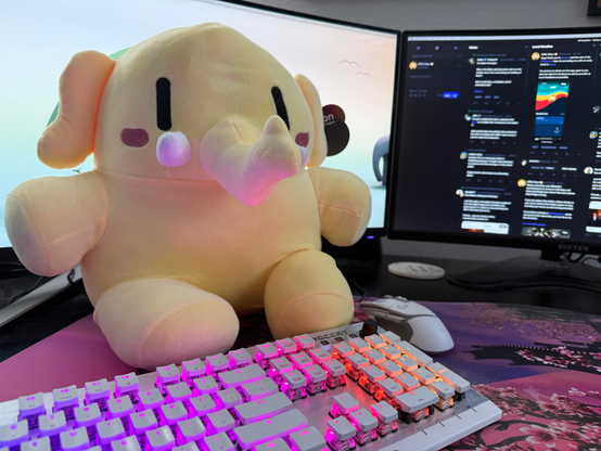 A photo I took of the plush Mastodon stuffed animal that arrived. #Plushtodon is the hashtag used for these photos and it's even on the tag on the ear. 

You can see part of my keyboard and two monitors in the photo along with my mouse.