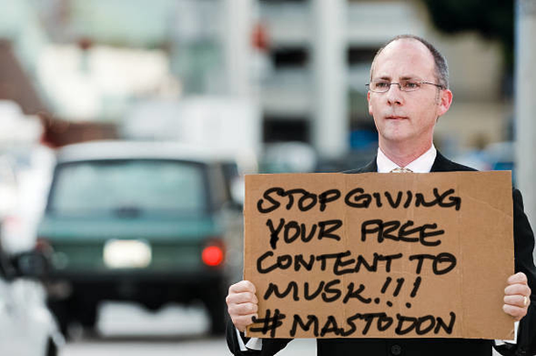 Man in a suit holding a cardboard sign saying “Stop giving your free content to Musk!!! #Mastodon