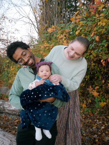 A black man and a white nonbinary person are leaning forward to either side to look at the baby that the man is holding. The adults are smiling and the baby looks curious.