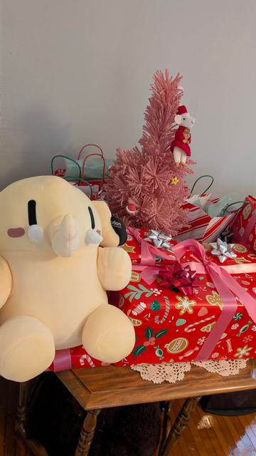 A picture of a yellow plush mastodon, the mascot of the mastodon project, is pictured in front of a small pink Christmas tree and several wrapped presents