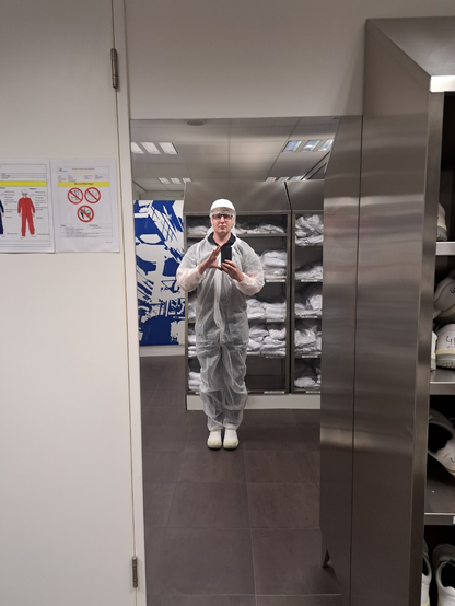 An awkward mirror selfie wearing a white disposable overall, hairnet, safety helmet and white safety shoes while standing in the dressing room in front of supplies of personal protective equipment.