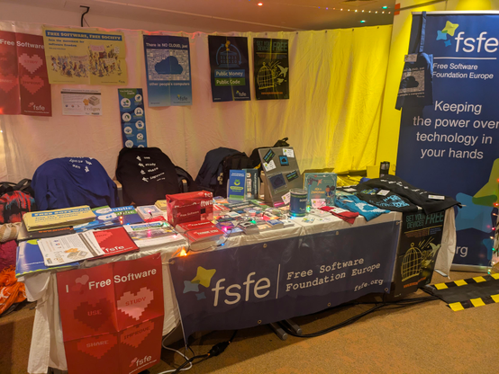 FSFE booth at the 38c3: two tables full of FSFE material. One table with different tshirts and a hoodie and the other with stickers, posters and info material 

This picture is a bit broader than the other one and you can also see FSFE posters hanging and and FSFE roll up