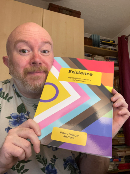 Peter, a cis white bald man holds up Existence, which has the intersex inclusive pride progress flag on the cover. 