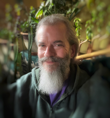 #selfie Middle-aged, white man with a graying full beard, mustache, and longish hair gently smiling for the camera, wearing a dark green hoodie and a purple t-shirt underneath, sitting at home in front of green houseplants.