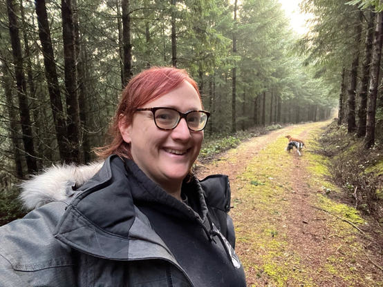 Nikki (@Aminorjourney) with her dogs on an early winter walk. She is wearing a thick winter coat with a fuzzy hood, has red hair brushed back, and thick-rimmed glasses. She is on a long forestry trail with tall trees on either side, while  a border collie and Labrador play in the distance. 