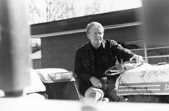 Jimmy Carter, sitting in the driver's seat of a tractor, dressed casually, looks contemplatively out into the distance. A utility building and winter trees without leaves can be seen in the background. 