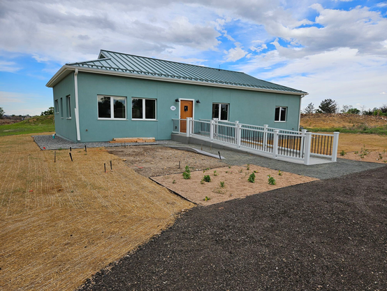 The new house complete. A green house with a green metal roof and white trim under a dramatic sky. There is a ramp with railings leading to the front door and preliminary landscaping has been done, with beds of new perennials, and grass seed under protective mats.