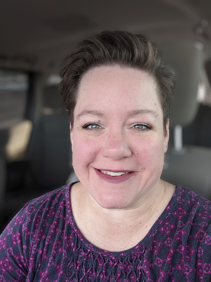 Selfie of a woman with short hair, sparkly green eyes, and a big smile, wearing a navy and fuchsia shirt. 