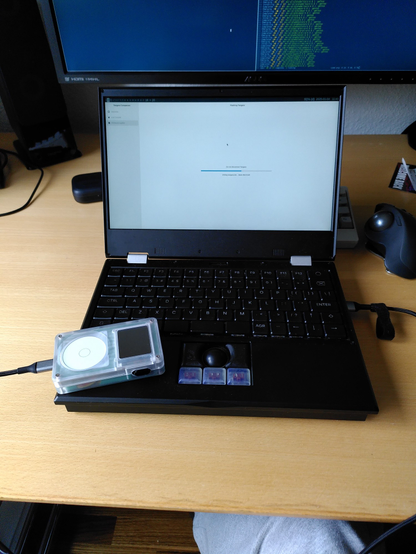 MNT reform laptop showing a progress bar. A tangara media player is placed on the palm rest of the laptop. The tangara's screen is off, it is connected to the laptop via USB cable.