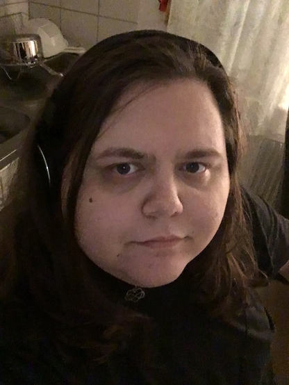 Fat trans girl sitting in kitchen, wearing headphones. Brown hair, blue eyes, wearing a choker or necklace with a heart and infinity symbol on it