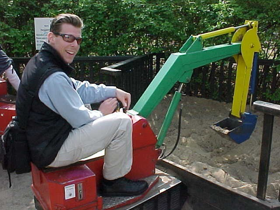 Picture of the author, a grown man, sitting on a coin-operated motorized backhoe digging sand in a playground.