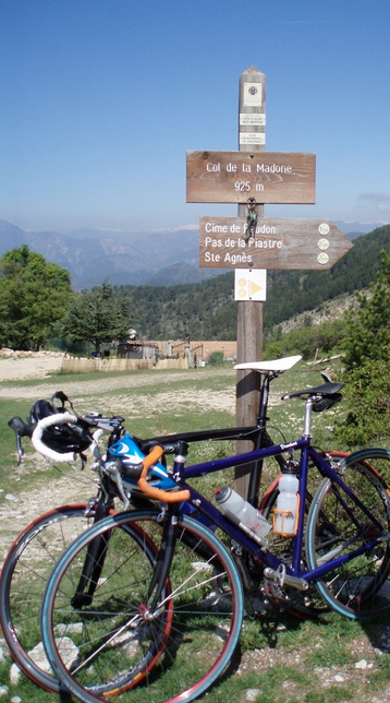 Two racing bicycles leaned against the wooden post with col sign for the Col de la Madone (925 m) near Nice, France. The bike in front is darkish blue with orange bar tape, red wheels, green tyres, a black saddle and two transparent water bottles. My surname is on the top tube near the seat post.