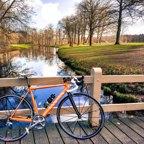 A bright orange Canyon racing bicycle leaned against a wooden bridge over a winding canal on Groeneveld estate in Baarn, Netherlands. The bike has white bar tape, white pedals, a white saddle and one black bottle. It has a Shimano Ultegra group but Campagnolo Eurus wheels. It is a sunny day with reflections of a few clouds in the water. The trees are bare but the grass is green.