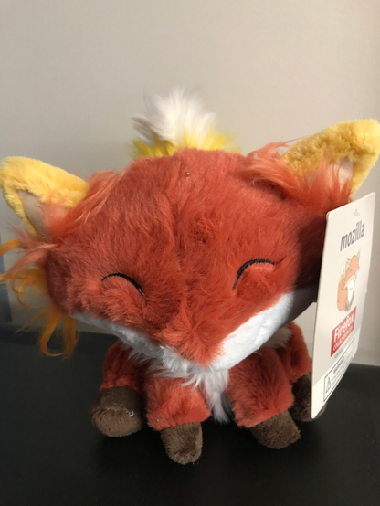 You can see a fox cuddly toy on a brown background (probably a table) and by a white wall. The fox has both eyes squinted (which makes him very cute). He is red and has white fur on his neck. His paws are brows. His legs and the part of the tail, which you can see, are yellow. On his right without hangs a shield. You can see the text "mozilla" and "Firefox", as well as his as drawing.