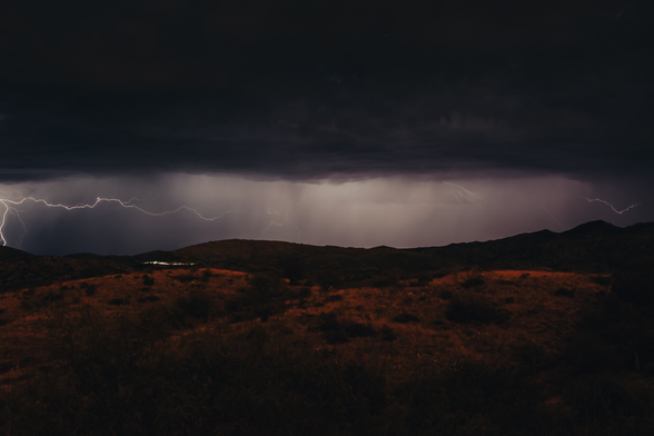 A desert landscape at night with lightening streaking across the sky. The clouds are illuminated and the ground is brown dotted with green shrubs and trees 