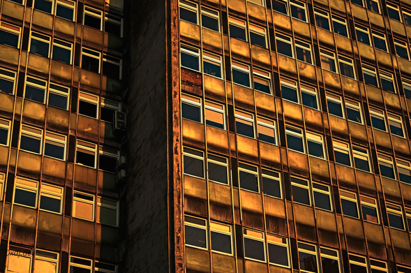 A facade of an old office building, with many identical windows, some of them partially covered by window blinds. The building has two parts, one part is set slightly back from the other, more visible in the photo. The concrete parts of the facade are visibly old and have some defects. The colour palette of the photo is warm, orange-yellow-ish, reflecting the golden hour during which it was shot.