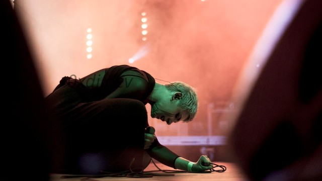 A person with short light blonde hair, in a long black dress with some see-through elements on its back, during a live music performance, kneeling on a stage between speakers/stage monitors. She’s holding a microphone. The makeup on her face imitates tears, it’s long dark stripes on her cheeks. The colours are warm with intense stage lights and stage fog behind the singer.