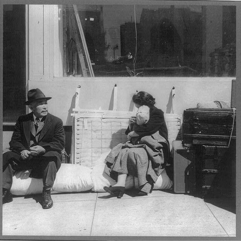 The image is a black and white photograph depicting three individuals sitting outside on what appears to be an urban street. On the left, there's a man wearing a dark suit with a hat, seated on a folded-up bench or structure with his hands clasped together in front of him as he gazes off into space.

In the center, another individual is reclining against a wooden slat-like surface that seems to be part of an outdoor seating area. This person appears older and wears dark clothing; they are resting their head on one hand while looking down with closed eyes or possibly asleep.

On the right side sits an elderly woman in what looks like a light-colored coat, seated beside some luggage consisting of suitcases stacked atop each other. She is wrapped in a shawl or blanket over her shoulders and holds another piece of clothing folded under her chin as she rests against the wooden slat surface with closed eyes.

The background shows part of an urban street scene including large glass windows reflecting buildings across from them, suggesting they are near commercial establishments like shops or offices. The overall ambiance seems somber, reflective, and evokes a sense of displacement or waiting for something to happen in their lives due to historical events such as World War II internment camps.

Additional context is provided by the caption which indicates this image was taken on April 1942 during evacuation proceedings involving [...]